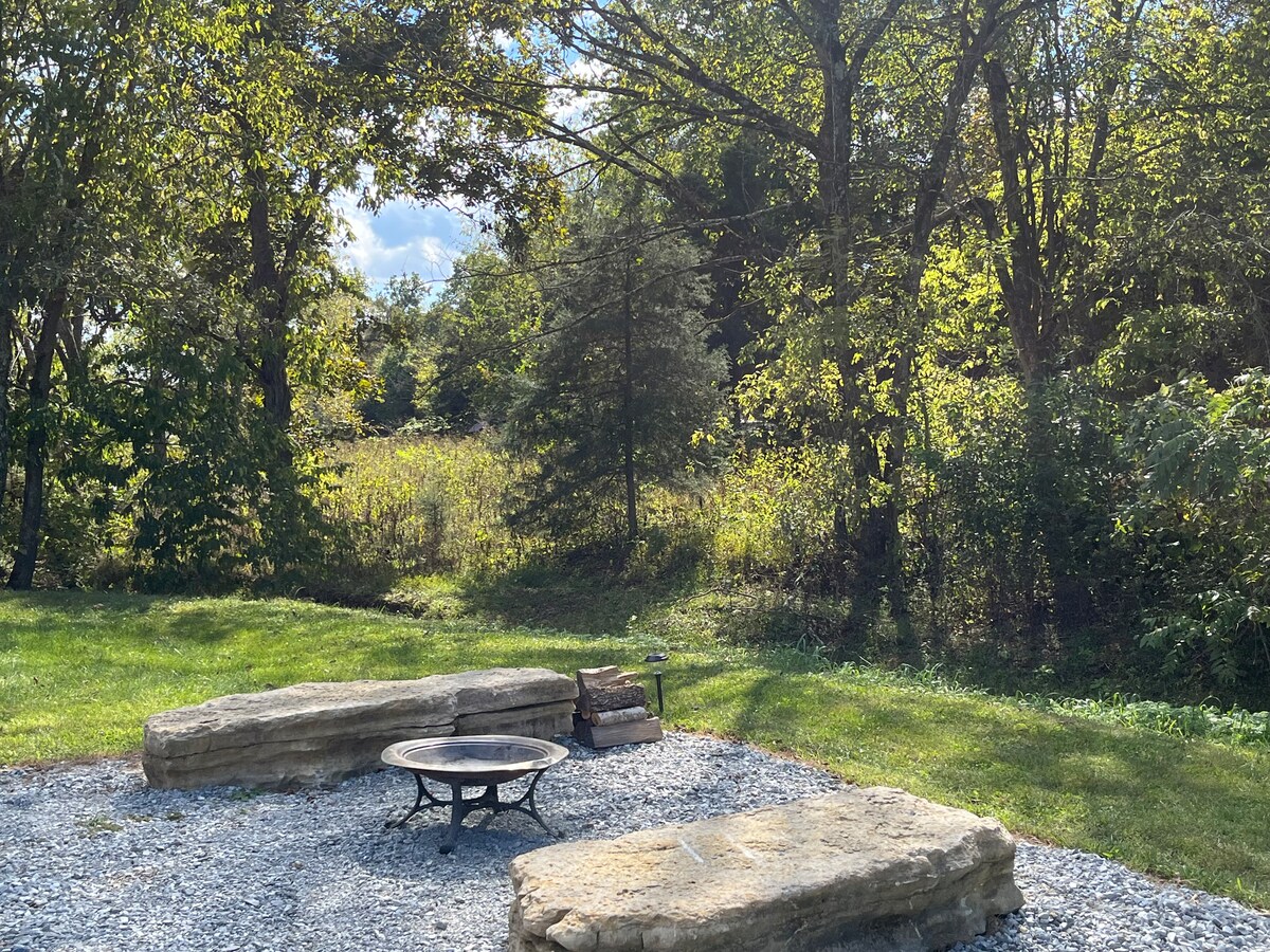 An outdoor seating area features two natural stone benches surrounding a fire pit. Lush greenery and trees provide a serene backdrop, with sunlight filtering through the leaves. The gravel ground enhances the rustic charm of this tranquil space suitable for relaxation and stargazing.