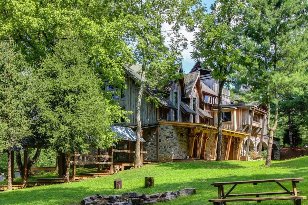 The Gatehouse is nestled among lush greenery, displaying a unique architectural style combining wood and stone elements. A spacious yard features picnic tables and stone fire pits, surrounded by trees. The residence's expansive porches extend outward, creating a welcoming atmosphere for outdoor gatherings.