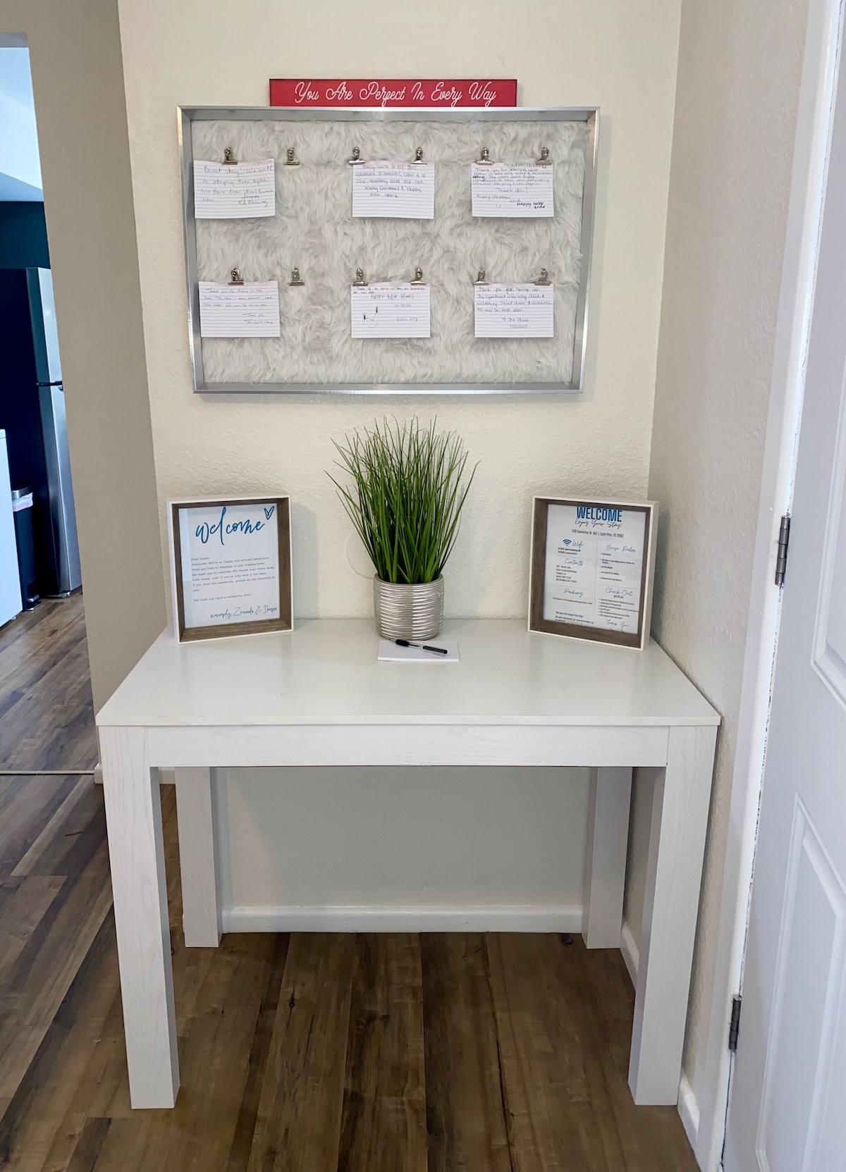 An entryway table is positioned against a light wall, complemented by a decorative plant in a metallic pot. A bulletin board above holds notes and welcome information. Two framed documents are displayed on the table, providing guests with useful details about their stay.