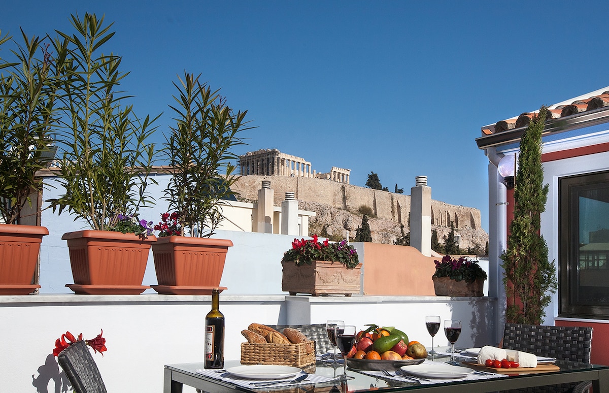 A private terrace is adorned with potted plants and a dining table set for a meal, featuring fresh fruits and wine. The Acropolis stands majestically in the background, providing a stunning backdrop against the clear blue sky.