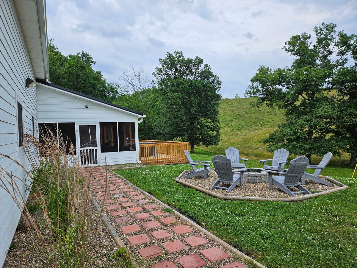 A spacious outdoor area features a circular stone fire pit surrounded by five Adirondack chairs. A pathway of red stones leads to a covered patio with screening. Lush greenery and rolling hills create a serene backdrop.