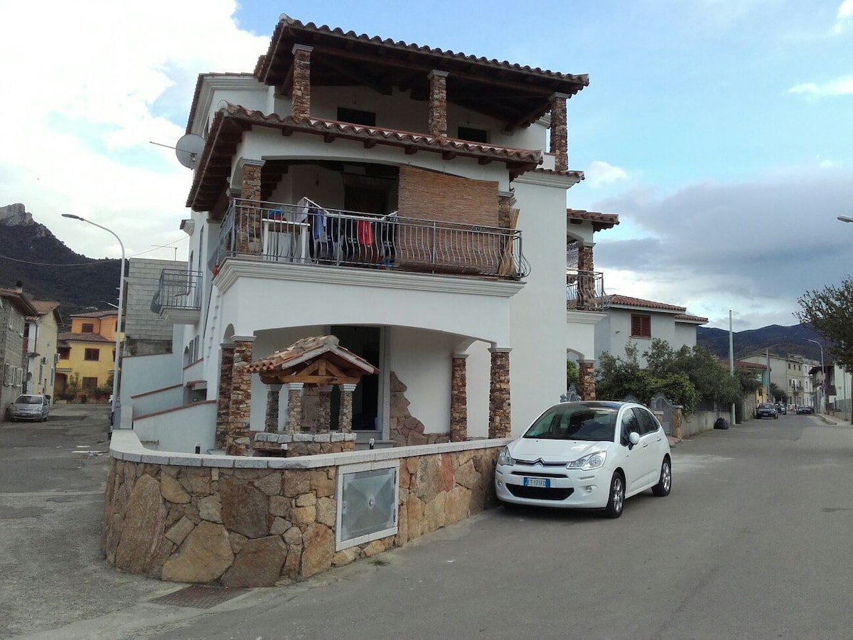 A charming three-story building is presented, featuring a combination of stone and white exterior. A spacious balcony with a decorative railing is visible on the upper floor. A small awning-covered area stands at the entrance, accompanied by a parked car on the street.