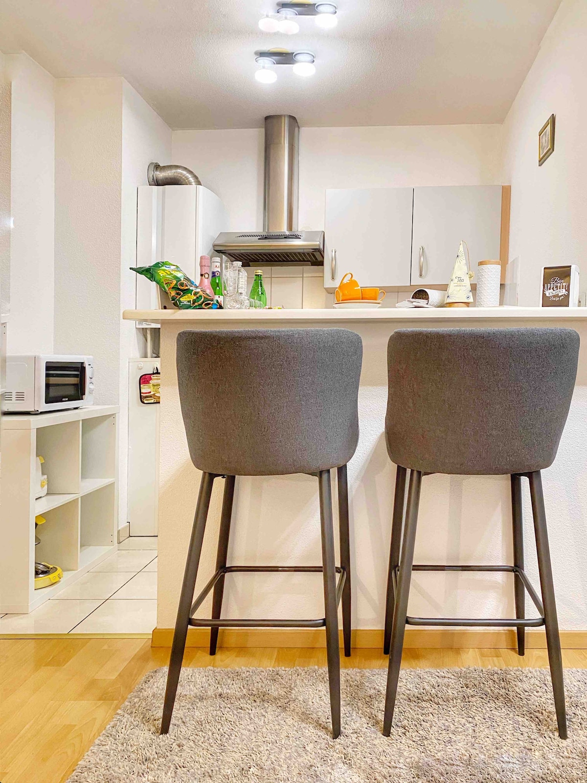 A kitchen area features a bar-style counter with two high, grey stools positioned on a light rug. A stainless-steel hood is visible above the counter, and kitchen appliances, including a microwave, are arranged in the background against a light-colored wall.