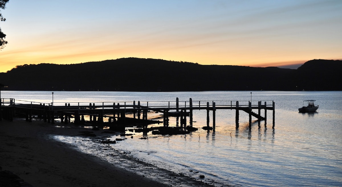 A serene waterfront scene is depicted, featuring a wooden pier extending into calm waters. Silhouetted against a vivid sunset, gentle hills rise in the background. The surface of the water reflects soft colors of pink and orange, creating a tranquil atmosphere.