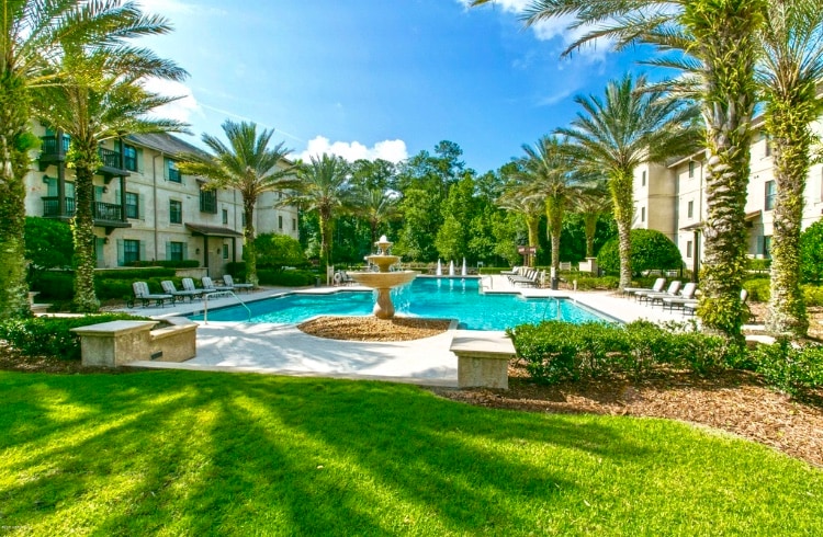 A well-maintained outdoor pool area is framed by palm trees and lush green landscaping. The sparkling blue pool features a central fountain and is surrounded by loungers, creating a serene environment for relaxation.