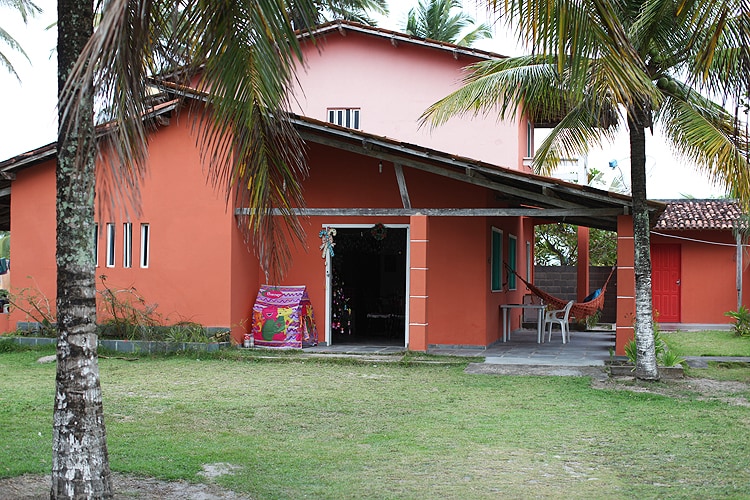 The house features a vibrant coral exterior, surrounded by palm trees. A shaded porch area includes a hammock and seating, providing a cozy outdoor space. The large lawn extends in front, offering ample space for leisure activities. A red door can be seen to the right.