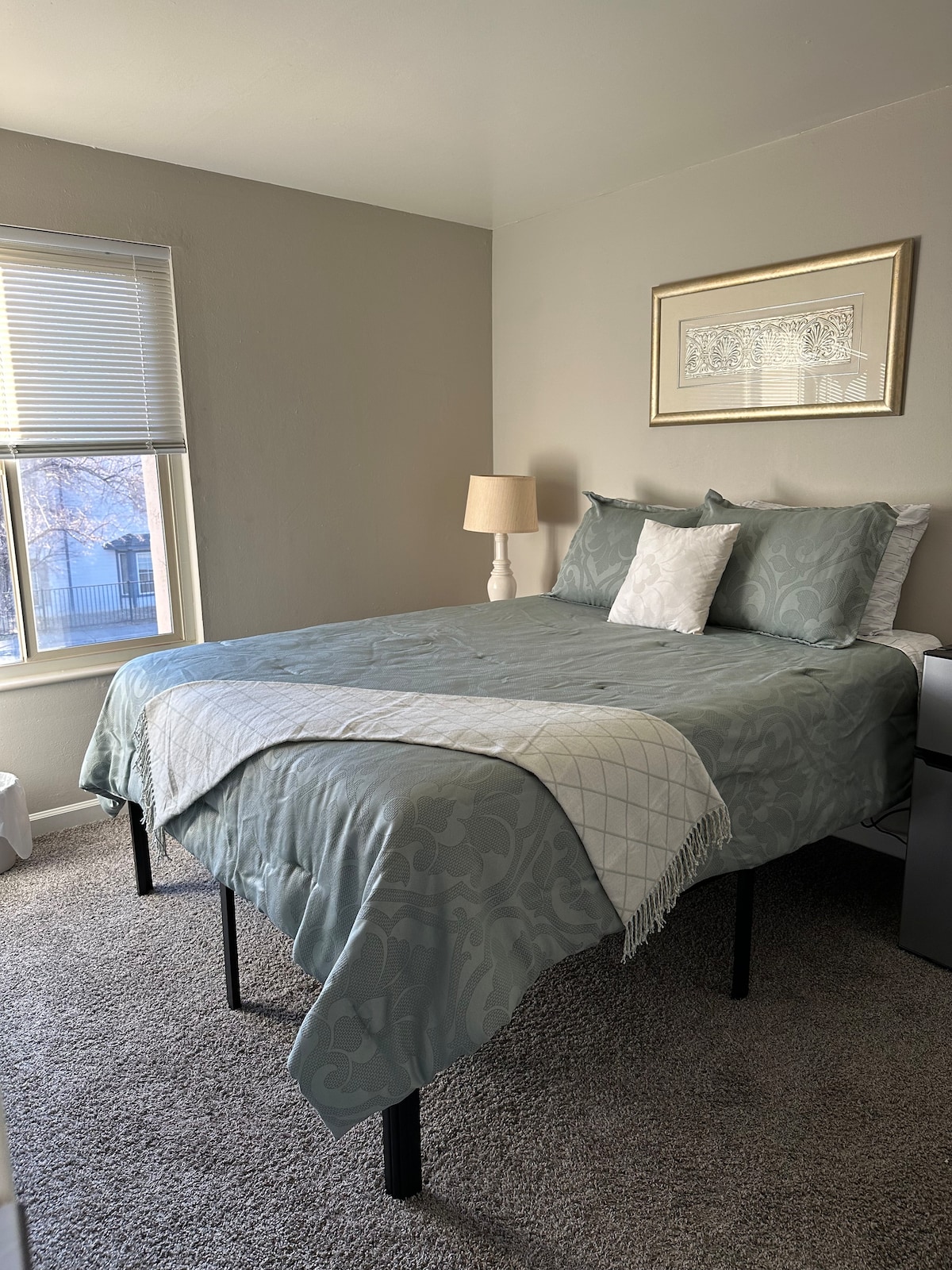 The room features a spacious bed, covered with a light blue bedspread and decorative throw. A bedside lamp stands on a black nightstand. Natural light enters through a window adorned with blinds, illuminating the neutral-toned walls and soft carpet.