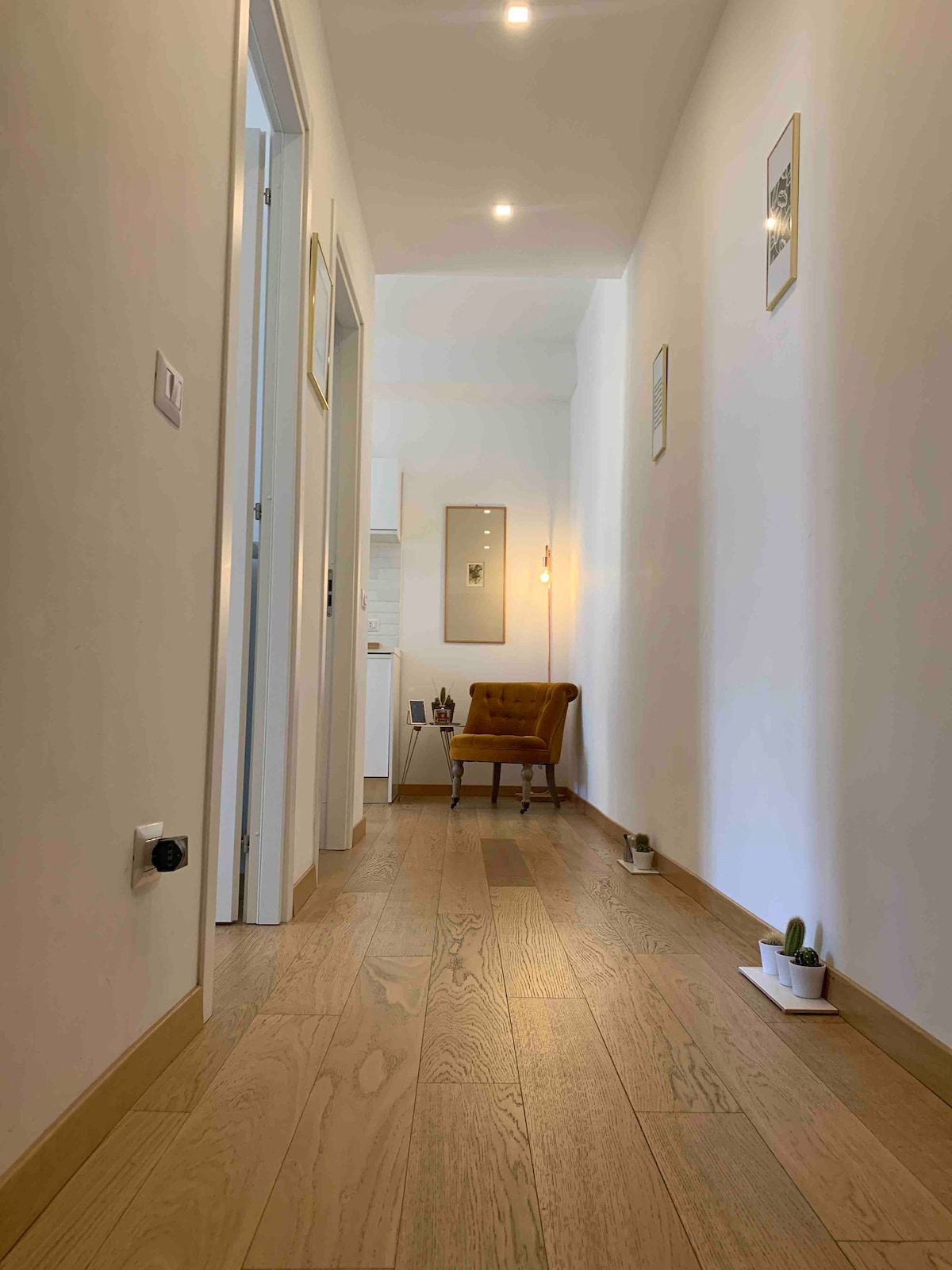 A welcoming hallway features smooth wooden flooring and white walls. A small chair is positioned against the wall, complemented by minimal wall decor and gentle lighting. Potted plants are placed along the floor, enhancing the serene atmosphere of the space.