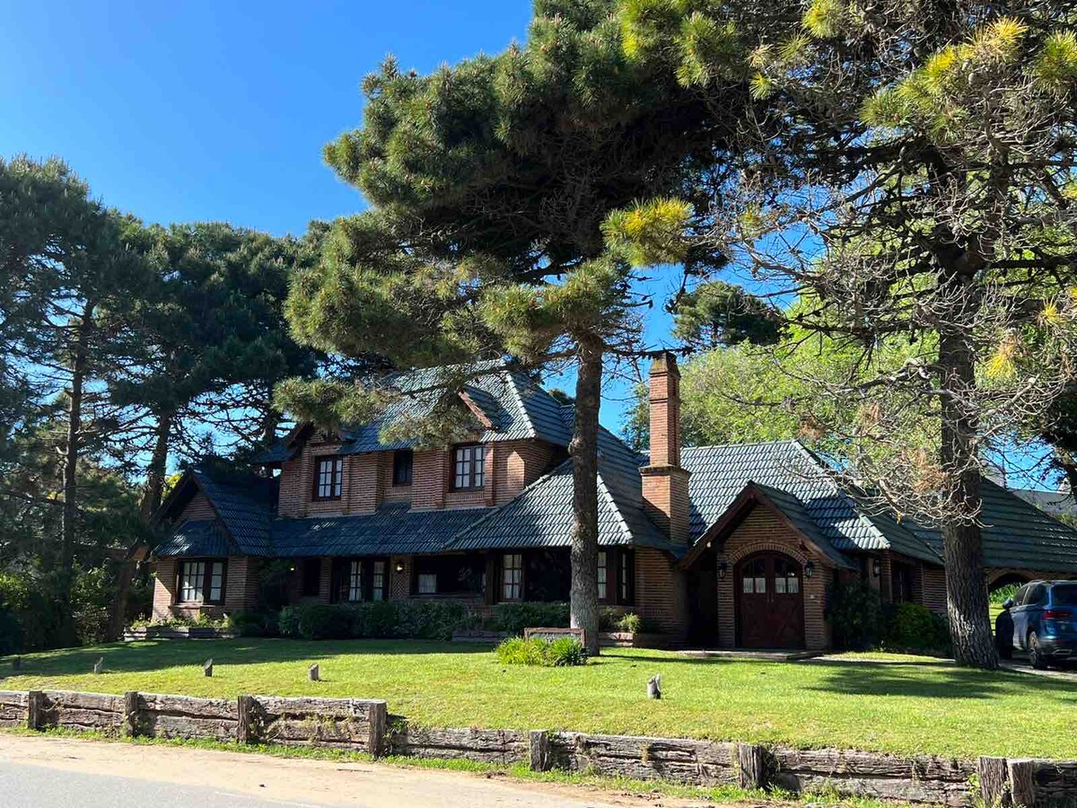 A spacious multi-level house is set amidst tall pine trees, featuring a prominent brick façade and a steeply pitched roof. Large windows and a welcoming front entrance are visible. Lush greenery surrounds the property, offering a serene outdoor environment.