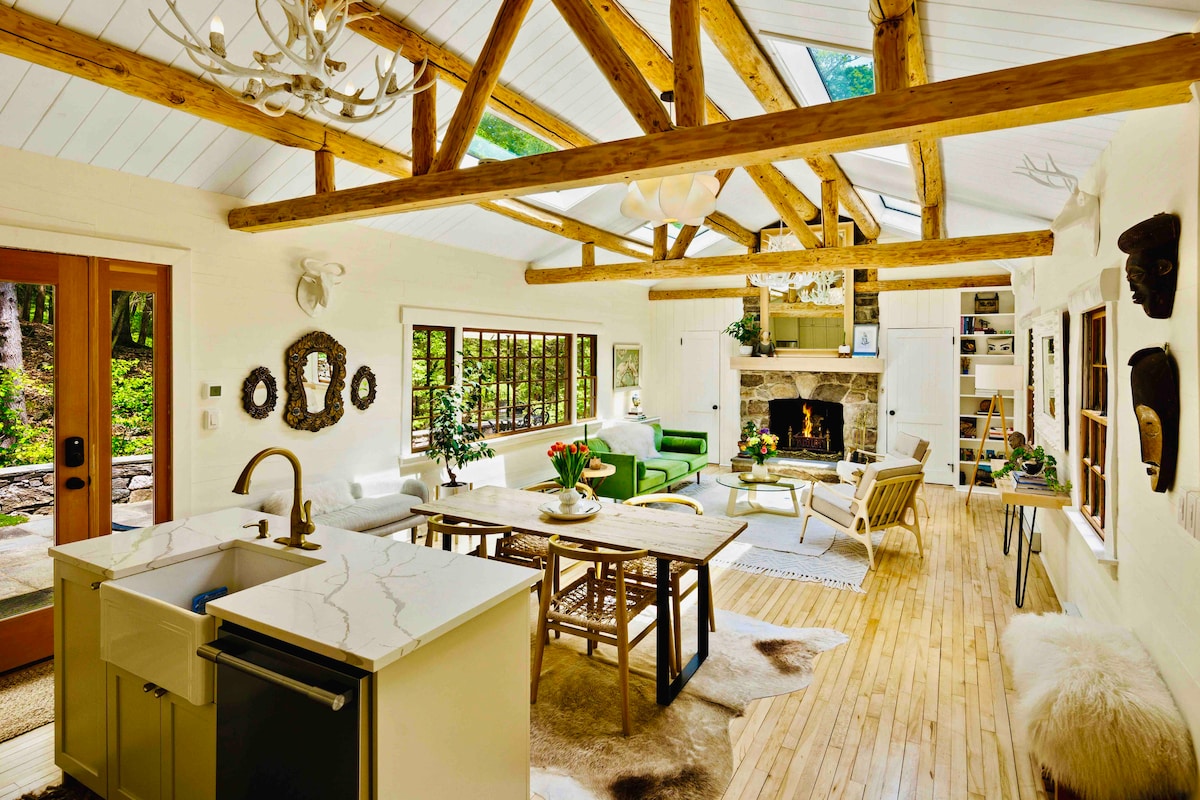 An open living space features a hand-scraped beam ceiling with skylights. A cozy seating area includes a green sofa and a coffee table surrounded by natural light. A fireplace built from local stones is positioned against a white wall, adding warmth to the room.