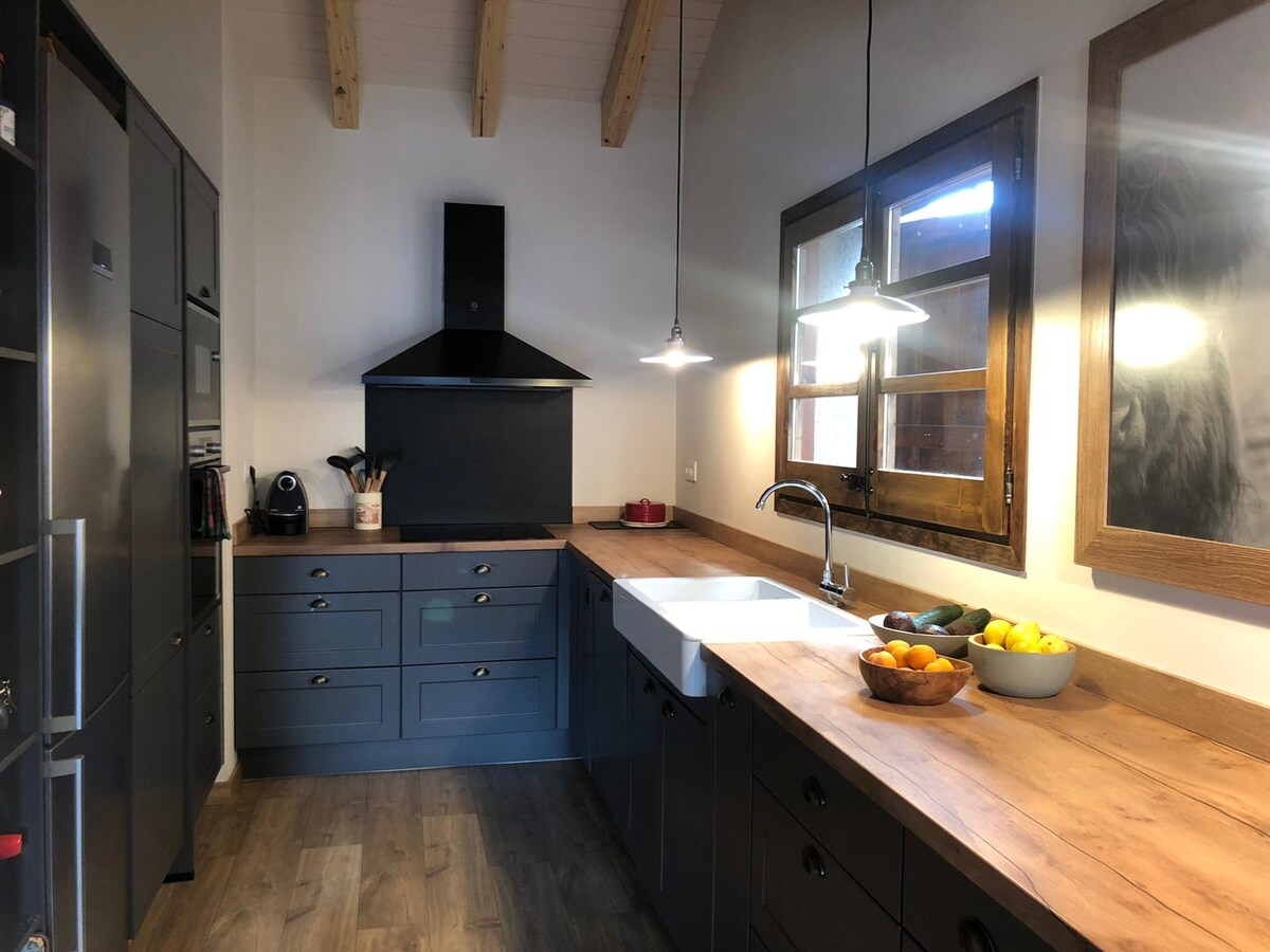 A modern kitchen features sleek gray cabinetry and a spacious wood countertop. A farmhouse sink is positioned under an adjacent window, allowing natural light to enhance the space. Fruit is arranged in a wooden bowl, and pendant lights create an inviting atmosphere.