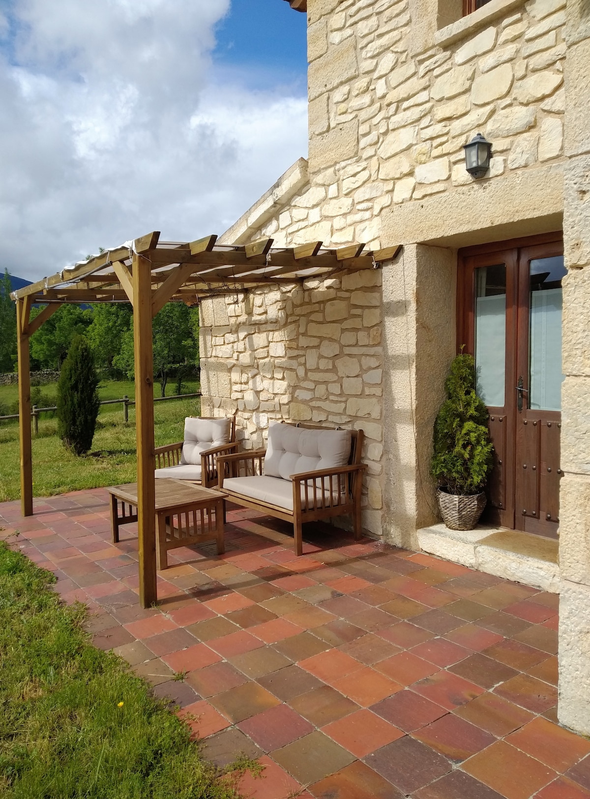 A covered patio area features two wooden seating arrangements with cushioned chairs. The rustic stone exterior complements the natural surroundings, which include green grass and shrubbery. A doorway with wooden accents leads into the casita, providing easy access to the outdoor space.