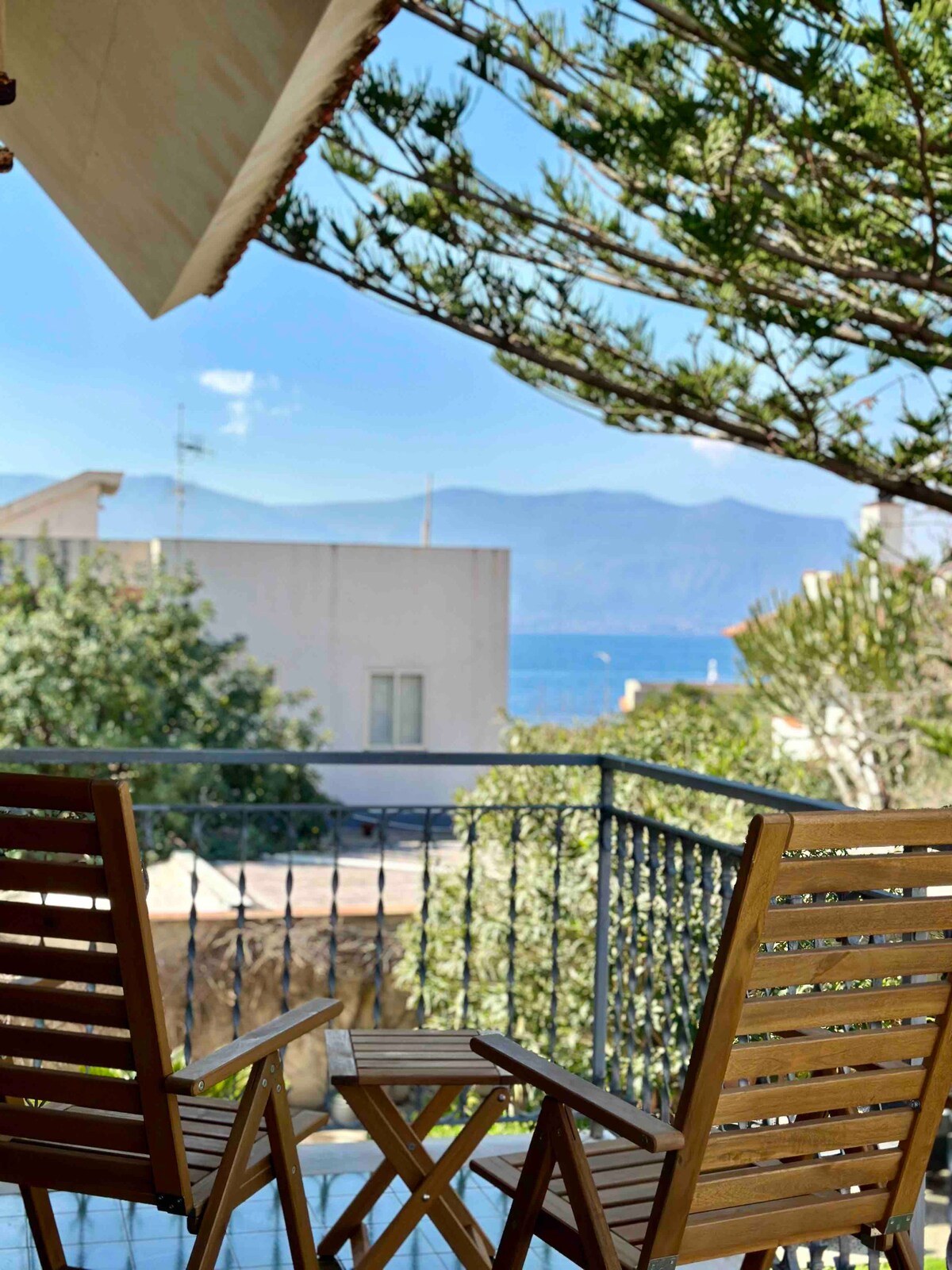 A private balcony is showcased with two wooden chairs arranged for relaxation. Scenic views of the sea and distant mountains are visible, framed by greenery. The space invites moments of calm and enjoyment of the surrounding natural beauty.