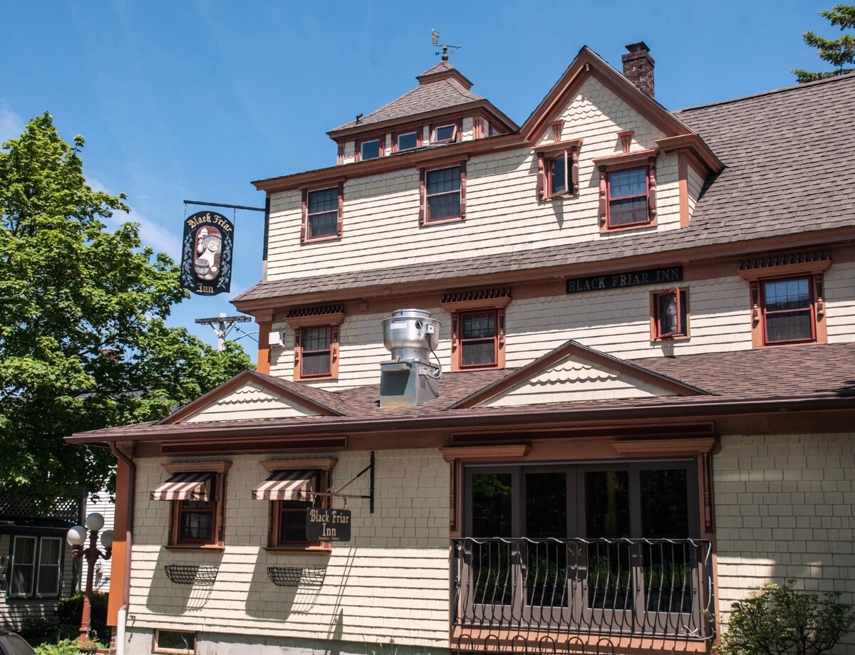 The Black Friar Inn and Pub displays a charming exterior featuring shingle siding and decorative details. The building includes multiple levels with gabled roofs, and a welcoming sign hangs above the entrance. Leafy trees surround the property, enhancing the inviting atmosphere.