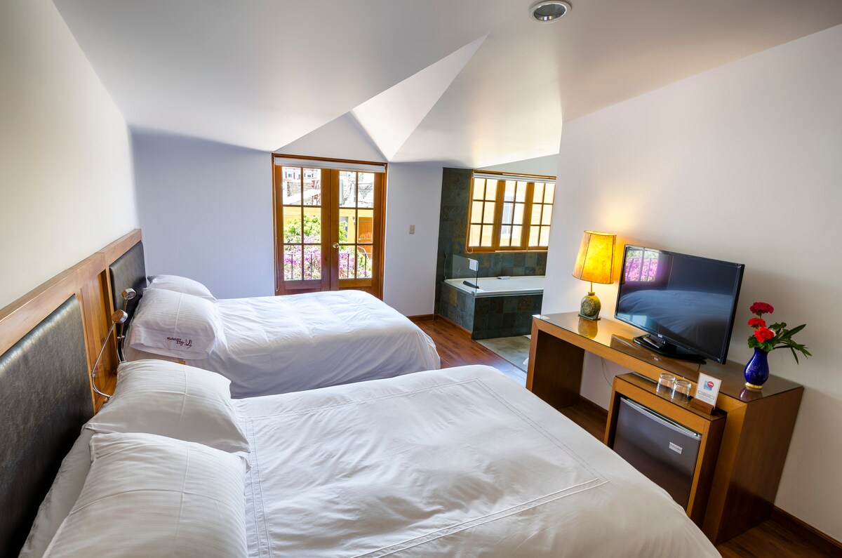 A well-lit room is furnished with two spacious beds, each adorned with white linens. A wooden desk holds a television and a floral arrangement. Large windows invite natural light and provide views of the outdoors, with a modern en-suite bathroom visible in the background.