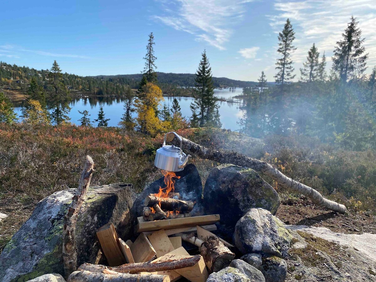 A fire pit is surrounded by stones, with flames visible as a kettle hangs above. The tranquil lake is reflected in the distance, framed by trees and autumn foliage. Smoke rises gently into the clear sky, contributing to a serene outdoor atmosphere.