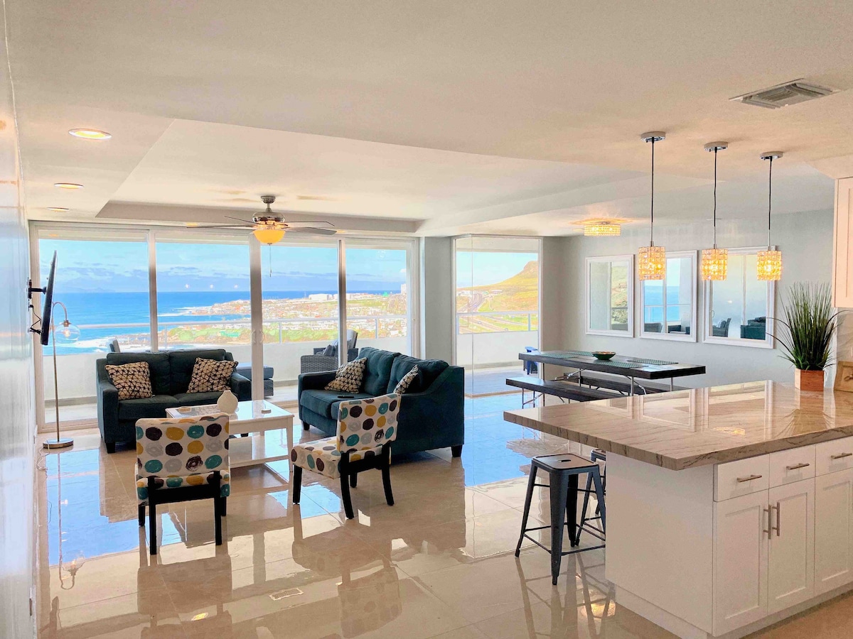 An open living space features large windows offering ocean views. A comfortable seating area includes a sofa and two patterned chairs around a coffee table. A dining table with seating is positioned near the kitchen, where sleek countertops and modern fixtures are visible.