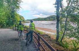 Two individuals wearing yellow safety vests are seen cycling along a pathway beside a river. Lush green trees provide shade on either side, and the water reflects the cloudy sky above. The distant landscape features rolling hills in the background.