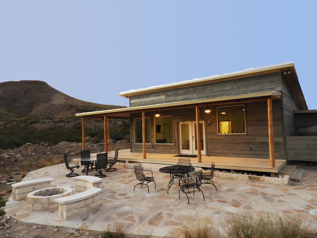 The modern cabin features a covered porch with seating options, including a dining table and chairs. A stone patio surrounds a central fire ring, perfect for gathering. The natural landscape and nearby mountain backdrop create a peaceful environment.