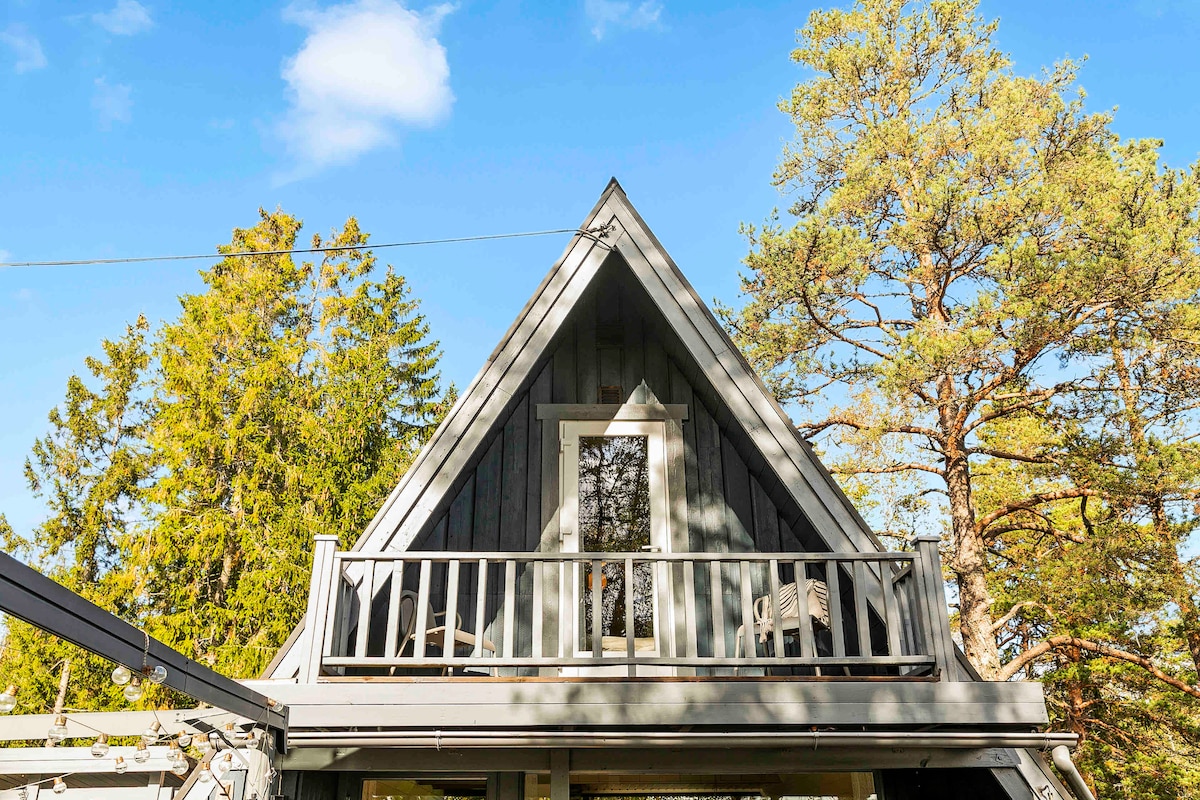 A charming A-frame cabin is depicted, framed by tall trees under a clear blue sky. A small balcony with a railing is visible on the upper level, providing an inviting spot for relaxation.
