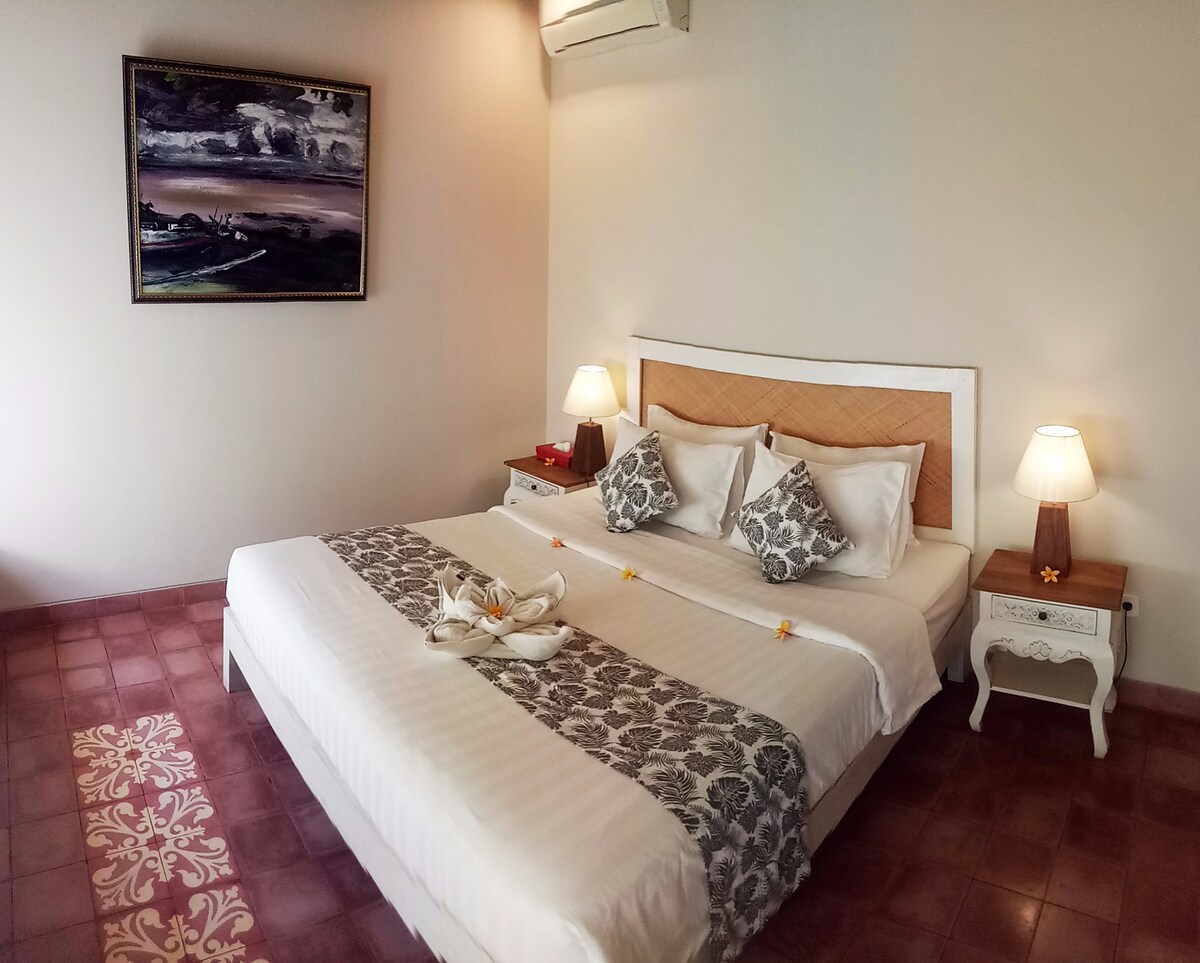 A spacious bedroom features a large bed with white linens and decorative throw pillows. Bedside tables are positioned on either side, each topped with a soft glowing lamp. A piece of artwork hangs on the wall, adding a touch of character to the room.