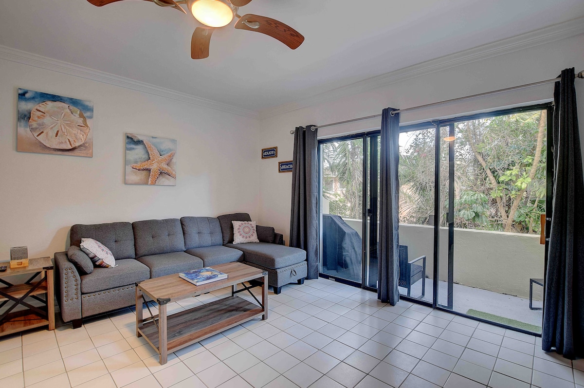 A comfortable living area features a gray sectional sofa, accompanied by a wooden coffee table. Large glass doors open to a balcony with views of greenery. Two dark curtains frame the doors, allowing natural light to brighten the space.