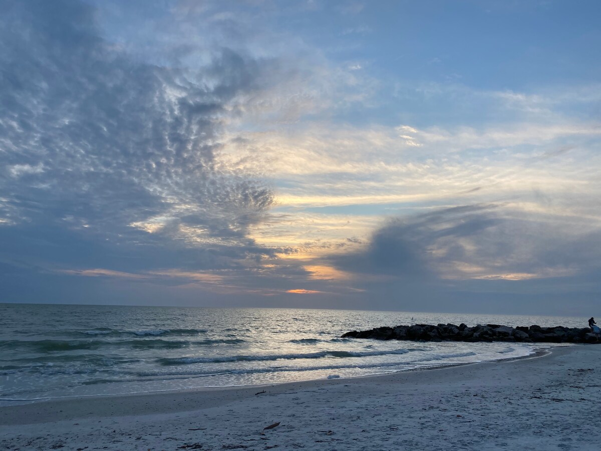 A serene view of the ocean horizon during sunset, with soft waves gently lapping at a sandy beach. Wispy clouds stretch across the sky, creating a gradient of colors as the sun sets beyond the water. Rocky formations are visible along the shoreline.