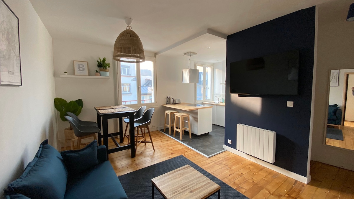 The spacious living area features a dark blue accent wall and a large television mounted on the wall. A cozy sofa is positioned next to a coffee table. A dining table with chairs is present, and the open kitchen is visible in the background, equipped with modern appliances.