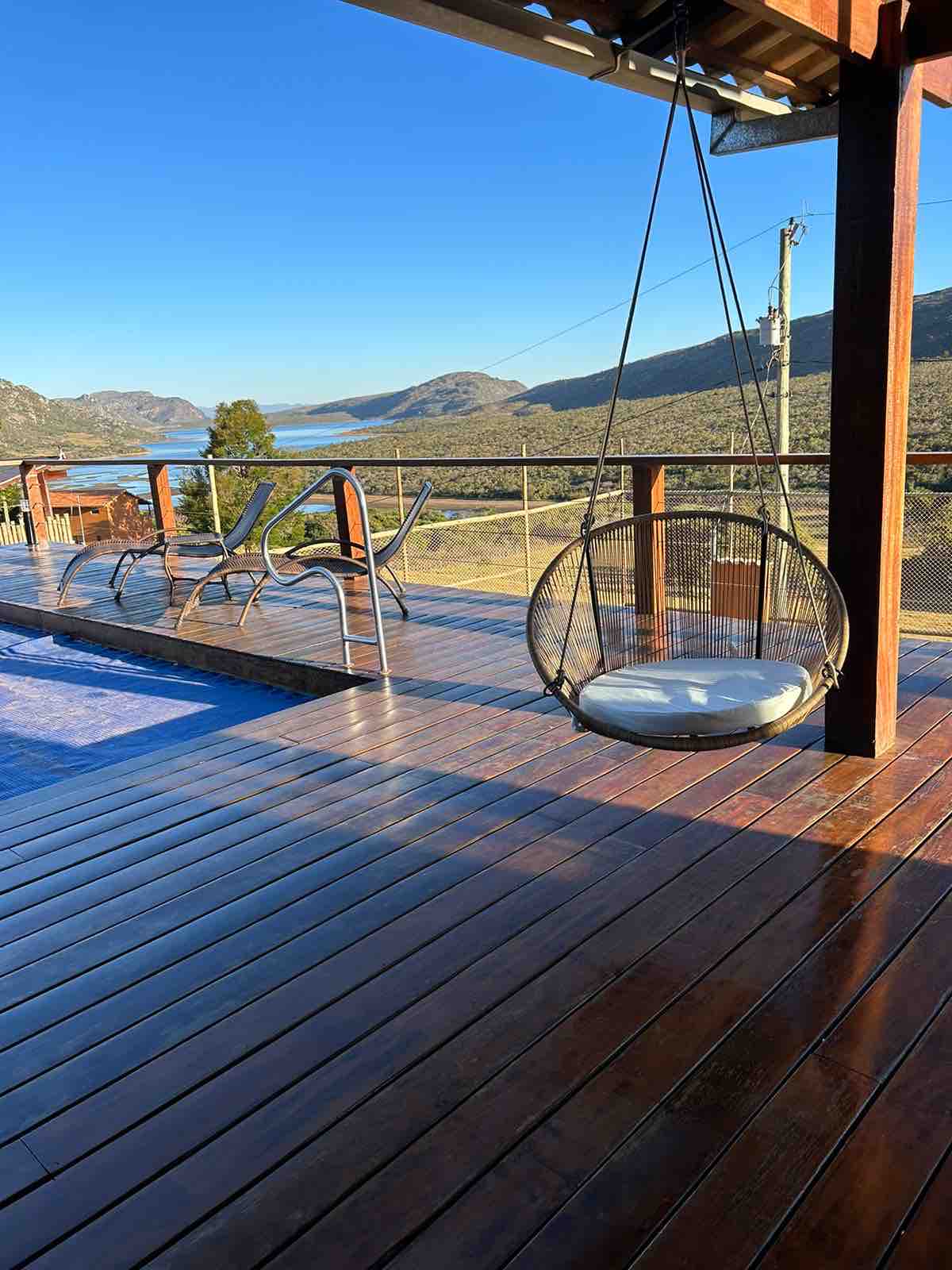 A spacious deck features a circular hanging chair with a cushion, surrounded by wooden flooring. Nearby, two lounge chairs are positioned beside a refreshing pool. Views of mountains and a serene landscape are visible in the distance under a clear blue sky.