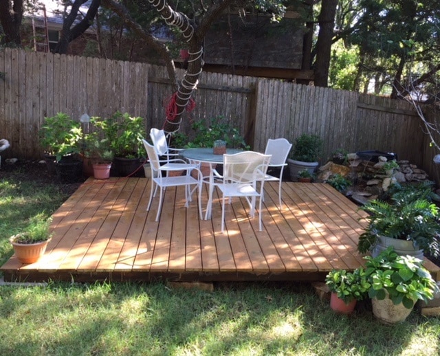 A wooden deck is seen surrounded by greenery, featuring two white chairs and a small table. Potted plants add a touch of nature, while a stone waterfall is visible in the background, enhancing the tranquil outdoor space.