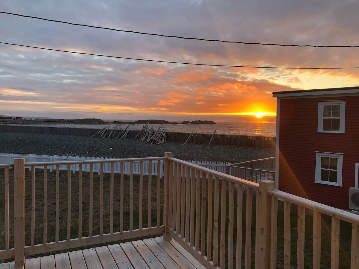 A serene sunset is captured over the Atlantic Ocean, displaying a gradient of oranges and yellows in the sky. A wooden deck offers a vantage point, and a breakwater of wooden posts extends into the calm water, enhancing the coastal view.