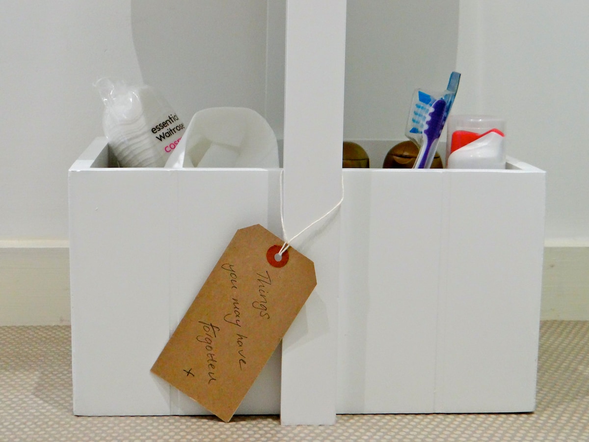 A white storage container is filled with various personal care items, including toiletries, toothbrushes, and other essentials. A brown tag is attached, inscribed with the message 'Things you may love together x.'