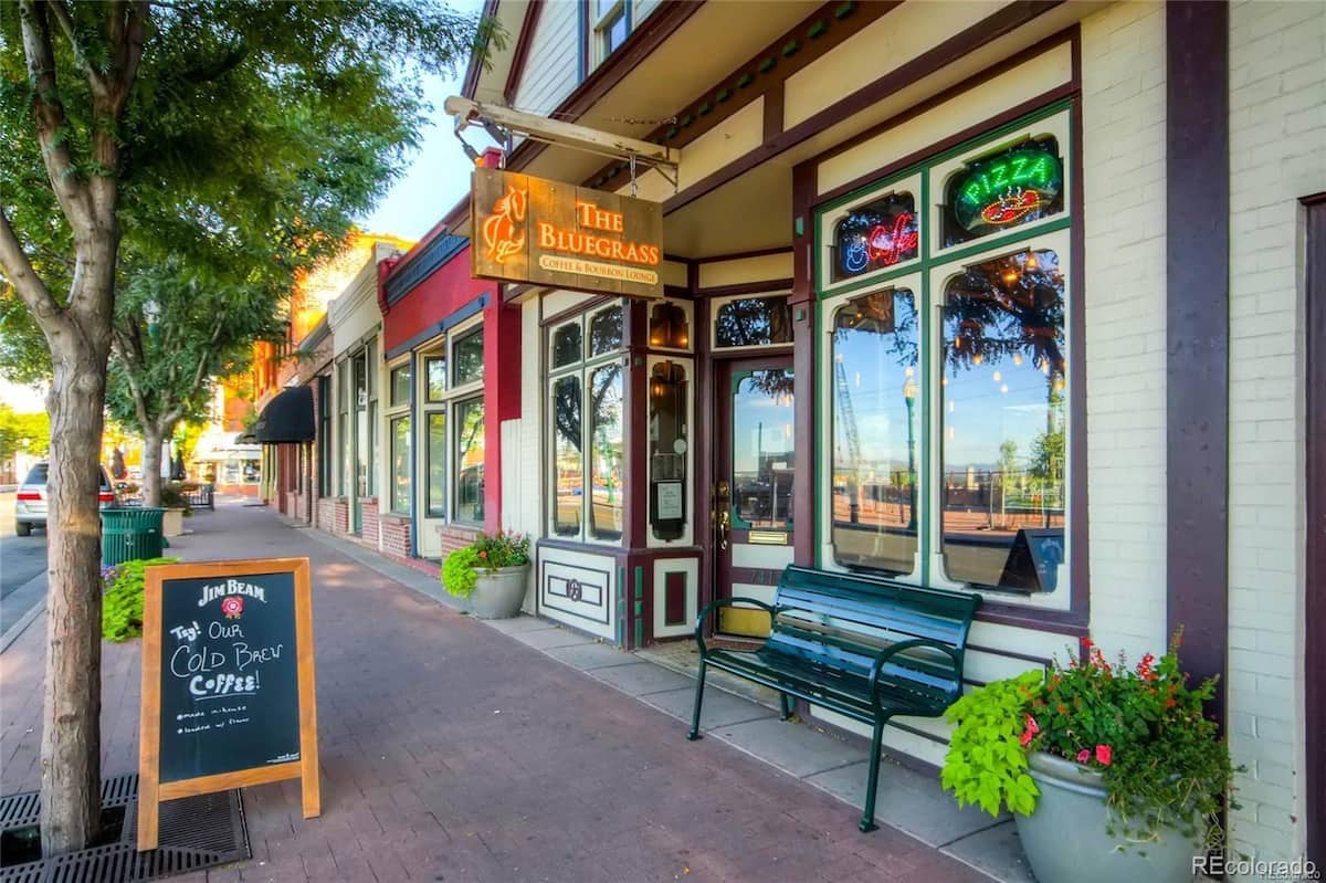 A welcoming storefront features large glass windows and colorful signage for 'The Bluegrass,' highlighting its offerings. Greenery from potted plants adds a natural touch, while a wooden bench invites passersby to sit. A chalkboard sign displays a message on the sidewalk, enhancing the neighborhood ambiance.