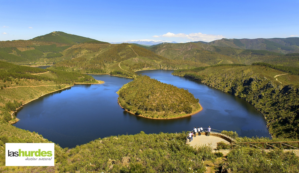 A wide view of a serene lake surrounded by rolling hills and lush greenery. The landscape showcases a curved shoreline, with a viewing platform that extends into the scene. Clear blue skies reflect on the water's surface, enhancing the tranquil setting.