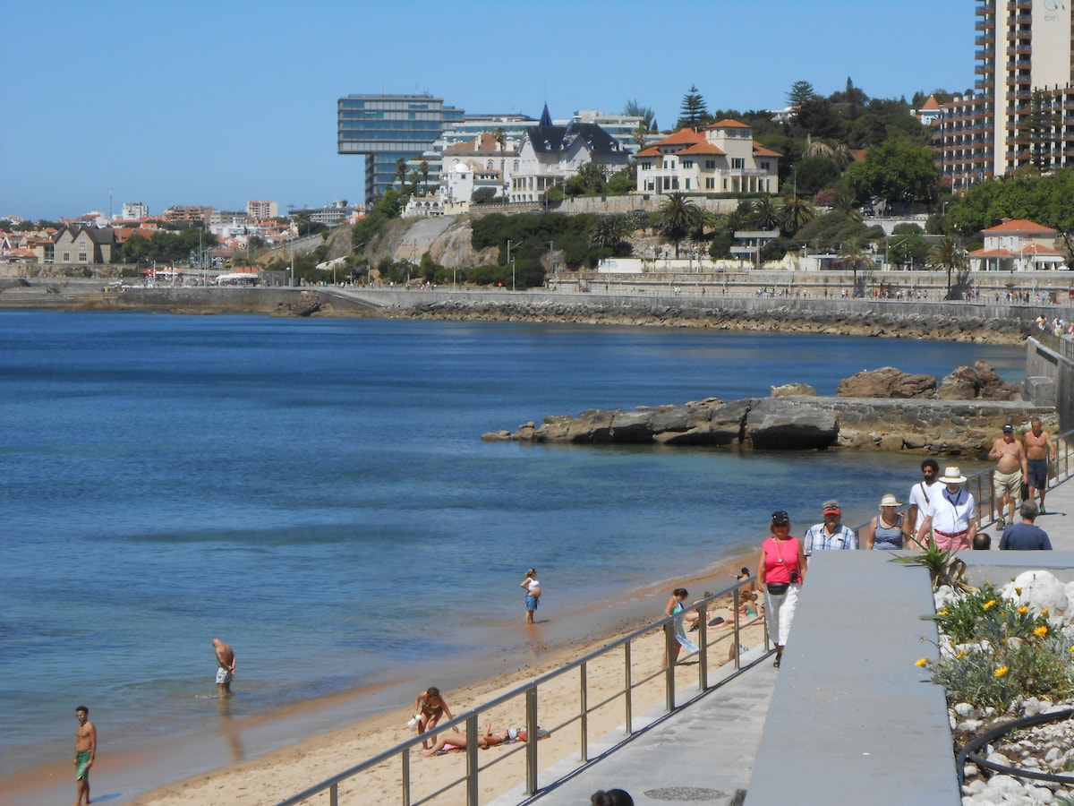 A sandy beach is shown with visitors enjoying the sun and water. Coastal buildings rise in the background, with colorful structures and greenery. A paved walkway runs parallel to the beach, providing access for individuals strolling or relaxing by the shoreline.