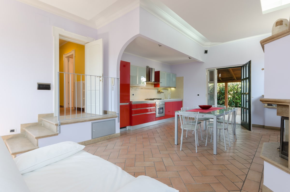 A welcoming open-plan living space showcases a modern kitchen with red cabinetry and stainless steel appliances. A dining table with white chairs sits adjacent to a comfortable sofa. Large glass doors provide access to an outdoor area, enhancing natural light in the room.