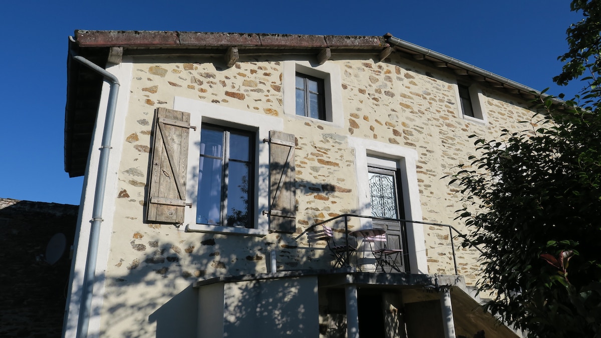 A stone building with rustic features is shown under clear blue skies. The exterior includes shuttered windows and a small balcony with a table and chairs, inviting an outdoor setting. Natural light highlights the texture of the stonework.
