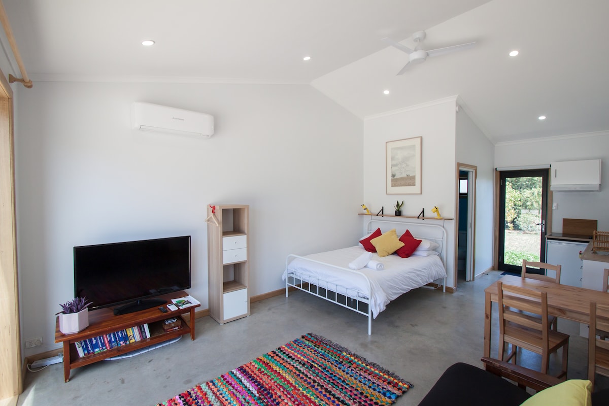 The studio features an open layout with a cozy queen bed dressed in white linens. A colorful rug complements the concrete floor. A television is positioned on a stand beside a small cabinet. A dining area is visible with wooden chairs around a table, and a kitchen area can be seen in the background.