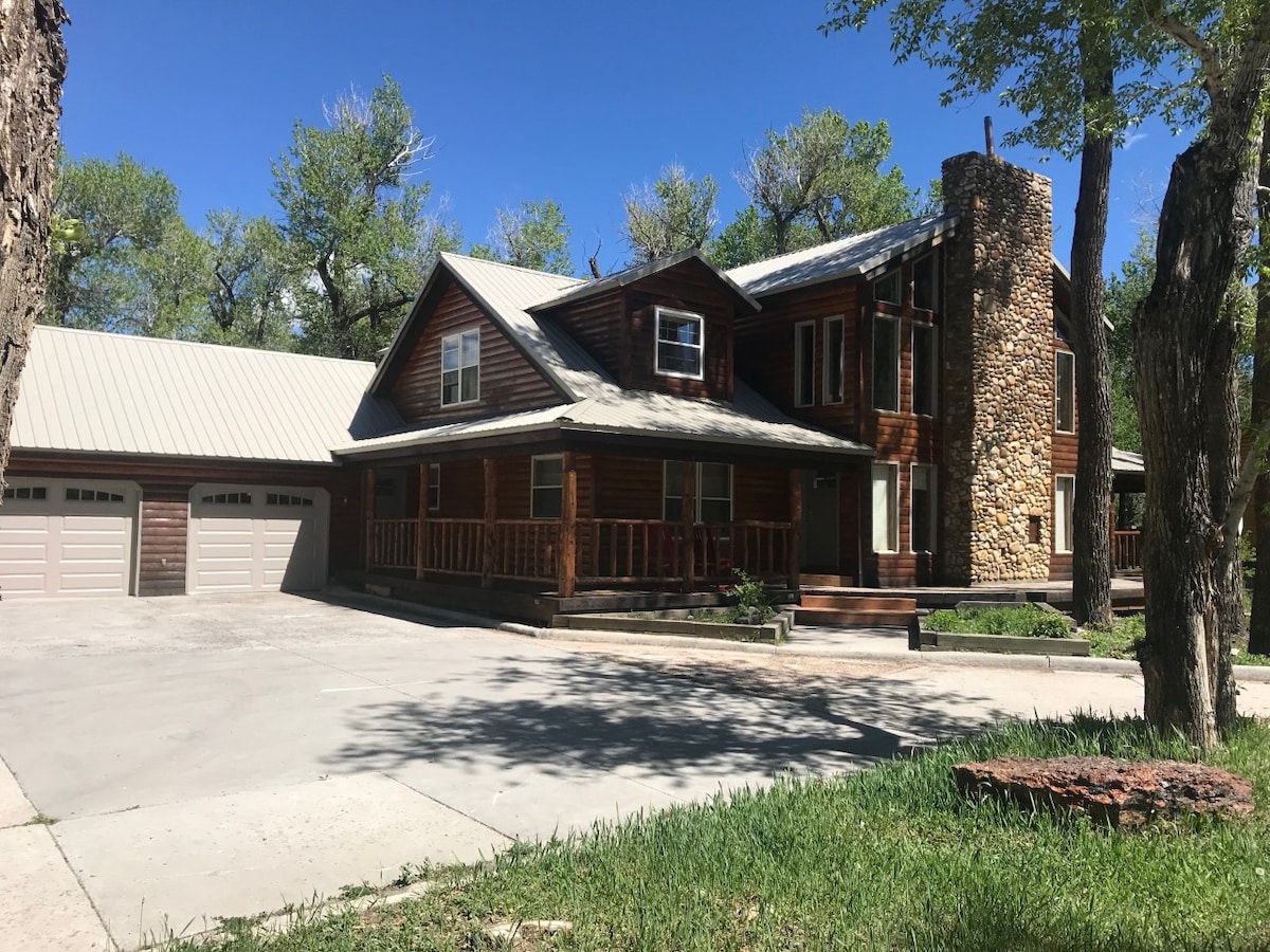 The rustic mountain home features a large, two-story wooden exterior with a stone chimney. A spacious driveway leads to a two-car garage, and the front porch is lined with wooden railings. Surrounding trees provide shade, enhancing the serene outdoor environment.