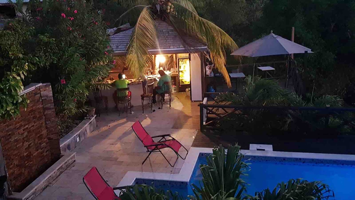 An outdoor courtyard area is shown, featuring a swimming pool surrounded by lush greenery. Two guests are seated at an outdoor kitchen bar, enjoying the evening ambiance. A dining area with an umbrella is visible along with two red lounge chairs, providing a relaxing spot near the pool.