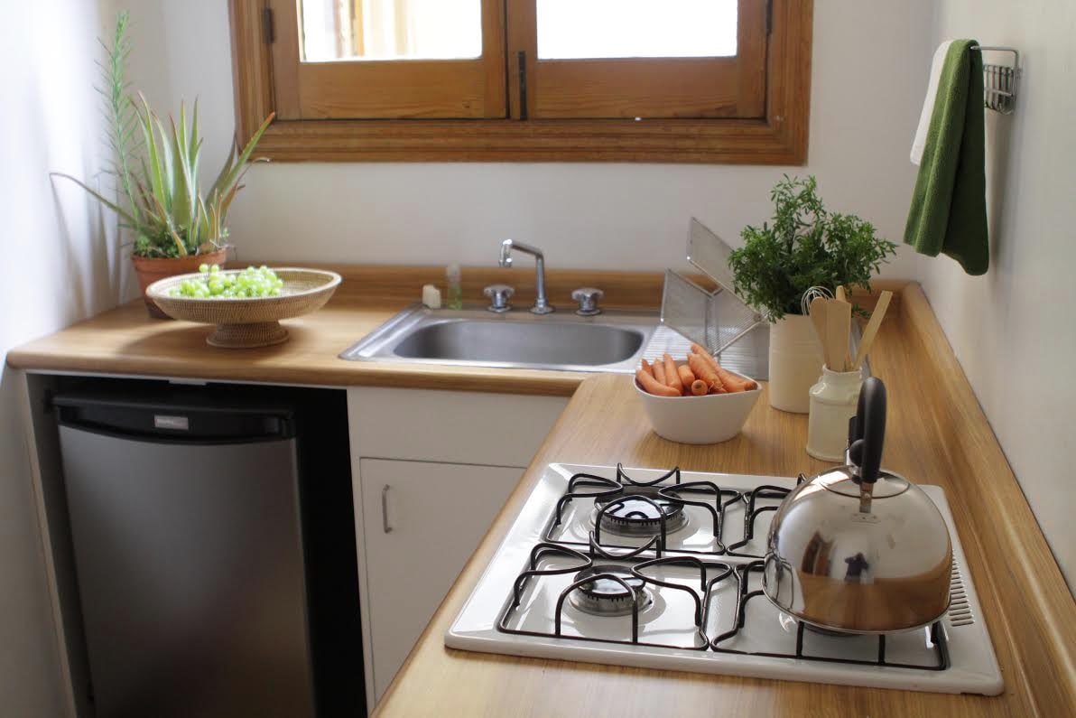 A well-equipped kitchen features wooden countertops and a stainless steel sink below a window. A small refrigerator is present, alongside a kettle on the stove. Fresh vegetables are arranged on a plate, while herbs are displayed in pots, adding a touch of greenery.