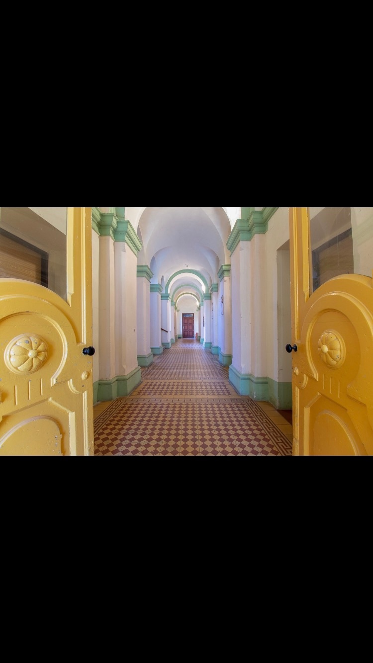 A bright corridor is illustrated, featuring arched ceilings and decorative details. The walls are painted in soft tones, with green accents contrasting against the light background. The corridor is lined with patterned tiles, leading to wooden doors at the far end.