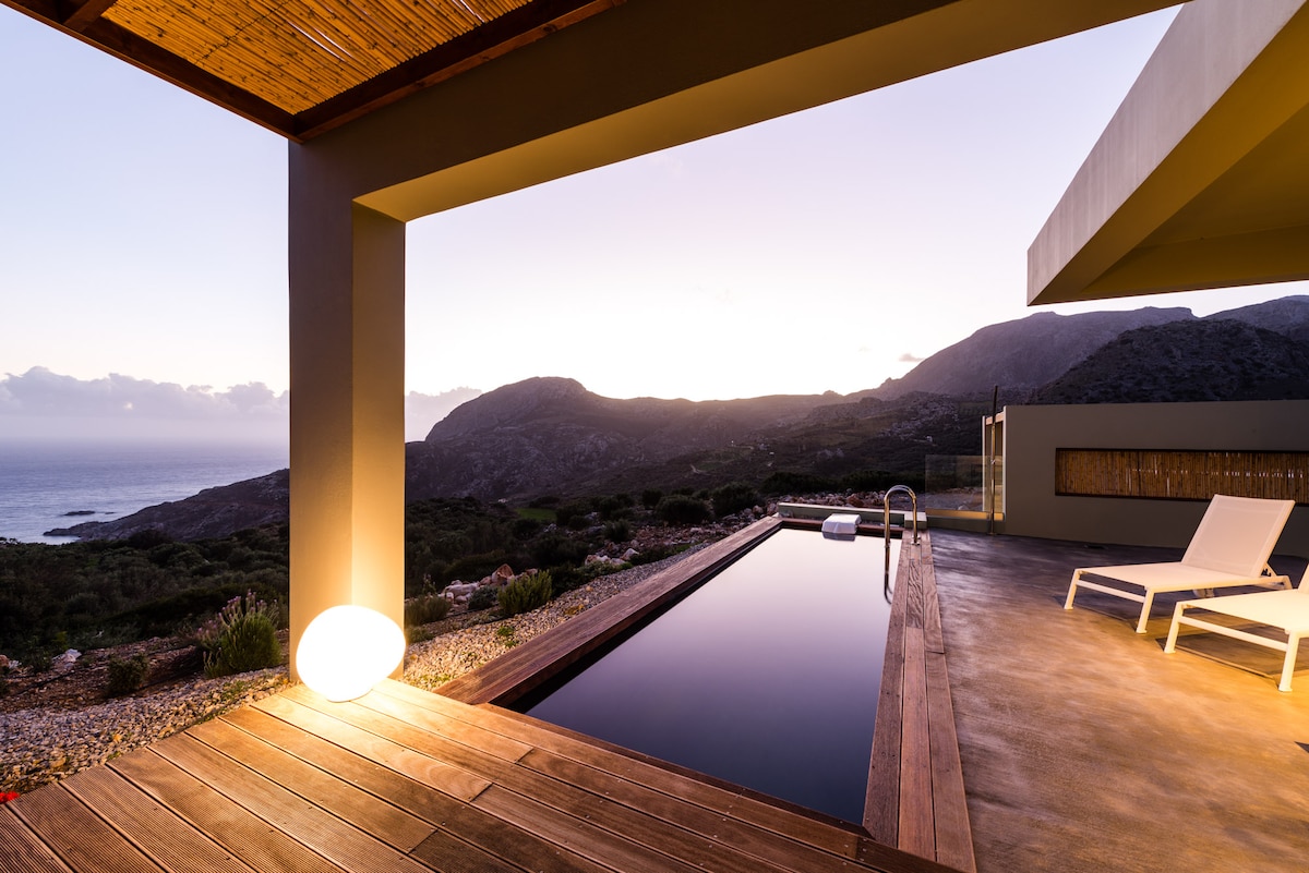 An outdoor terrace features a private pool with smooth wooden decking. Monochromatic lounge chairs provide a place to relax while overlooking the serene sea and mountainous landscape. Dusk light casts a soft glow, enhancing the tranquil ambiance of the setting.