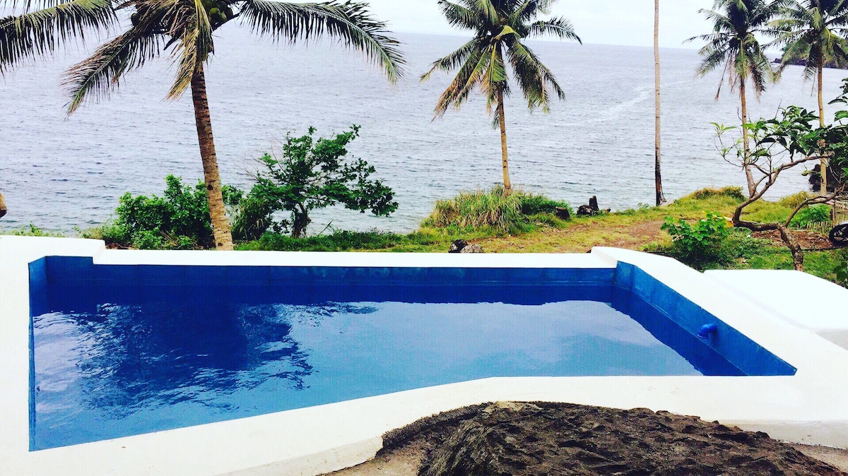 A serene private pool is framed by lush greenery and palm trees, with the Pacific Ocean visible in the background. The pool's clear blue water reflects the surrounding natural beauty, creating a peaceful retreat for relaxation.