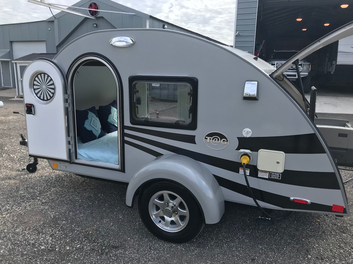 The exterior of a retro-style teardrop trailer is shown, featuring a streamlined design in grey with black accents. The door is open, revealing a cozy sleeping area inside, while the side includes a visible electrical outlet and a wheel hub with spokes.
