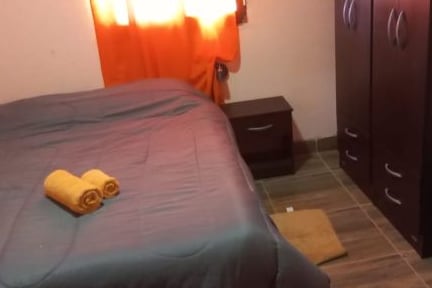 A spacious bed is adorned with neatly rolled towels, positioned beside a dark wood nightstand. Natural light filters through a vibrant orange curtain, while a small rug lies on the tiled floor near a dark wooden wardrobe with multiple drawers.