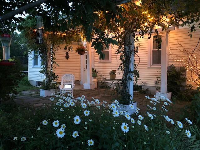 A serene outdoor patio space features white wicker seating nestled among blooming daisies. Soft lighting hangs overhead, illuminating the natural surroundings and creating a peaceful ambiance. The house's exterior, characterized by simple white siding, complements the inviting atmosphere.