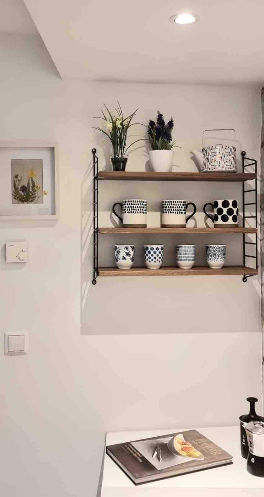 A wall-mounted shelf displays an assortment of decorative mugs in various patterns, accompanied by potted plants and a patterned pitcher. A framed artwork hangs nearby, contributing to a cozy kitchen ambiance.