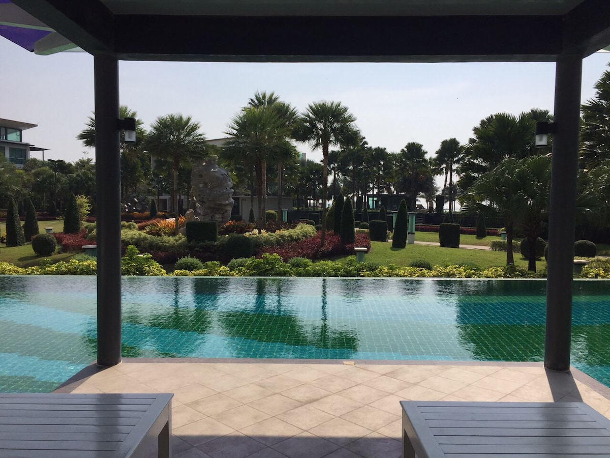 A clear view of a well-maintained garden is visible beyond a tranquil swimming pool. Lush greenery, palm trees, and vibrant flower beds surround the area, creating a serene atmosphere. Comfortable seating is positioned at the poolside, inviting relaxation and enjoyment of the scenery.
