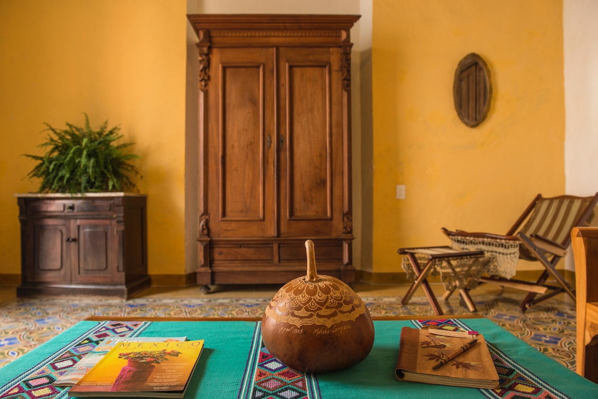A wooden armoire stands against a warm yellow wall, accompanied by a small decorative brown gourd on a vibrant green table. Two magazines are placed on the table, while a cozy armchair and a folding chair are visible in the background, enhancing the inviting interior.